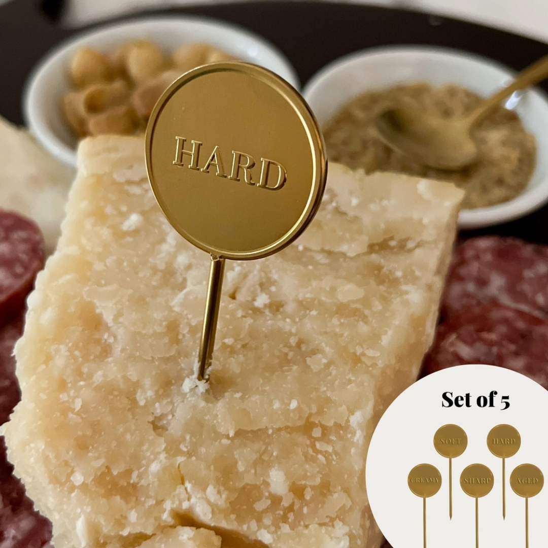 A close-up of a cheese board with a round gold metal pick engraved with the word "HARD" inserted into a wedge of hard cheese. Small bowls of nuts and spread are blurred in the background, and a circular inset at the bottom right shows five matching gold picks labeled as a set of 5.