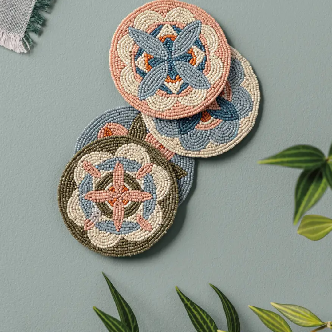 Three round woven coasters with intricate geometric mandala patterns in beige, blue, and terracotta, arranged on a muted sage green background with decorative green leaves.