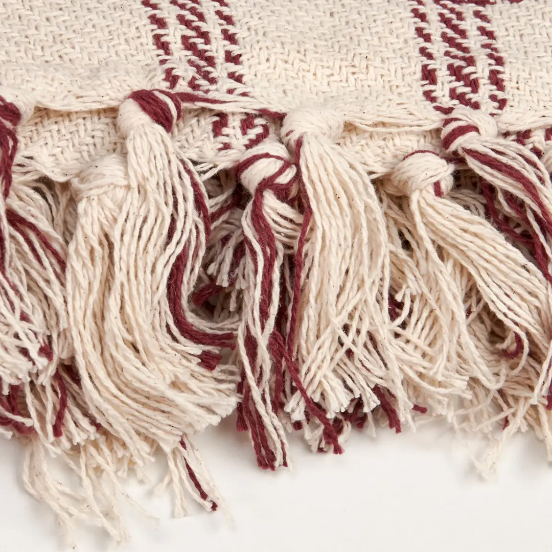 Close-up of a woven textile with cream and burgundy stripes, showing fringe tassels and part of a patterned woven surface.