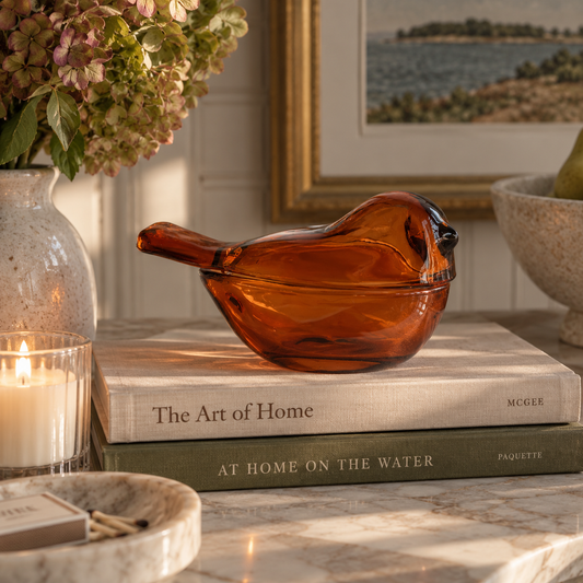 An amber-brown translucent glass bird figurine resting on stacked books on a marble surface, with a lit candle, vase of flowers, and framed wall art in a styled interior setting.