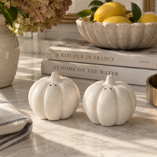 Two white ceramic pumpkin-shaped salt and pepper shakers on a marble countertop, styled with a striped cloth, a vase of flowers, stacked books, and a bowl of lemons in warm natural light.